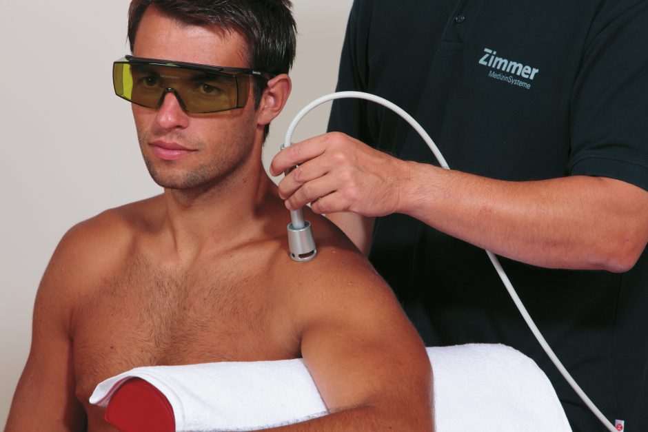Ein Mann sitz Oberkörperfrei und Schutzbrille. Der Therapeut steht dahinter und behandelt mittels Lasertherapie seine Schulter